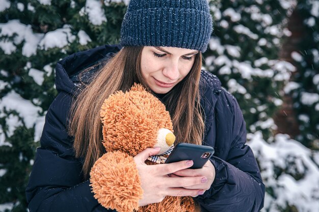 Una mujer joven sostiene un oso de peluche y un teléfono inteligente en sus manos en tiempo de nieve