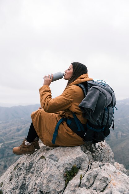 Mujer joven con su mochila sentada en la cima del pico de la montaña bebiendo el agua de una botella