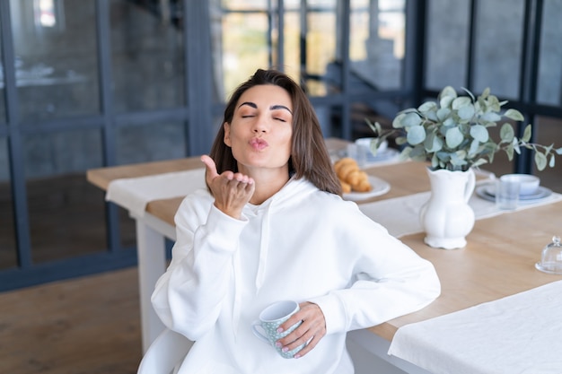 Foto gratuita mujer joven con una sudadera con capucha blanca cálida en casa en la cocina, comienza su día con una taza de café