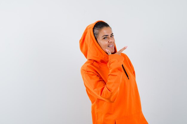 Mujer joven en sudadera con capucha naranja apuntando a la derecha con el dedo índice y mirando feliz