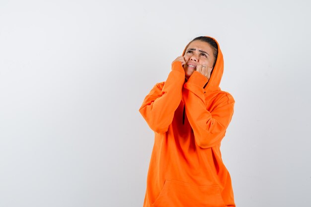 Mujer joven en sudadera con capucha naranja poniendo las manos en las mejillas, mirando hacia arriba y mirando acosado