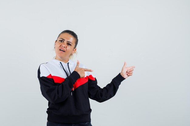 Mujer joven en sudadera colorida apuntando hacia atrás y mirando ansioso, vista frontal.