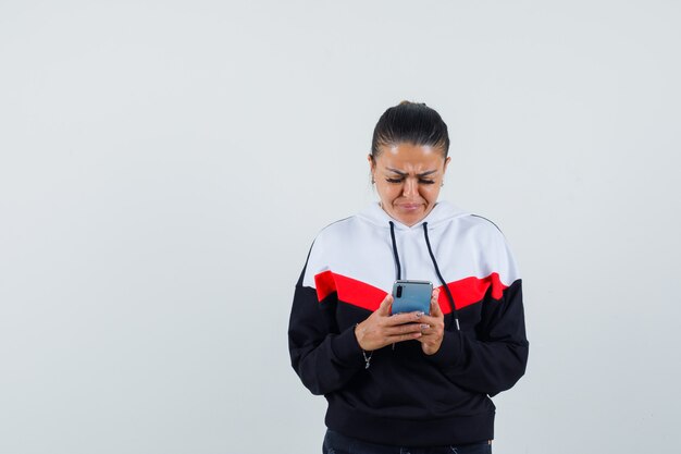 Mujer joven en sudadera colorida mirando el teléfono y mirando triste, vista frontal.