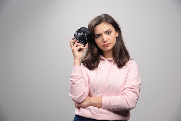 Mujer joven en sudadera rosa sosteniendo la cámara y mirando con enojo.