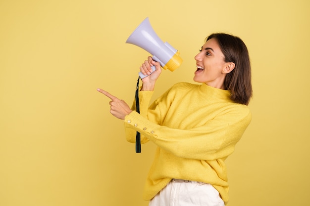 Mujer joven en suéter amarillo cálido con altavoz de megáfono gritando hacia el dedo índice apuntando hacia la izquierda