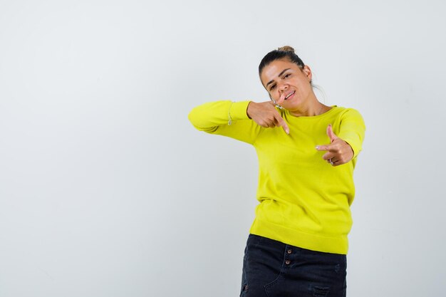 Mujer joven en suéter amarillo y pantalón negro apuntando a la cámara con los dedos índices y mirando feliz