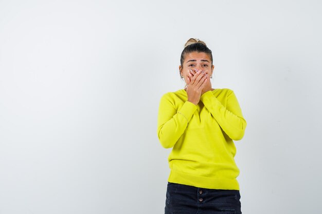 Mujer joven en suéter amarillo y pantalón negro cubriendo la boca con las manos y mirando sorprendido