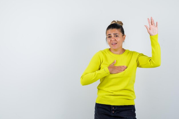 Mujer joven en suéter amarillo y pantalón negro levantando la mano y sosteniendo la mano en el pecho y mirando emocionado