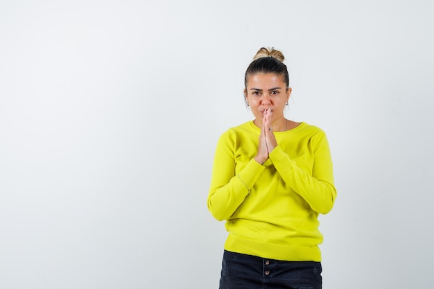 Mujer joven en suéter amarillo y pantalón negro mostrando gesto de namaste y mirando serio