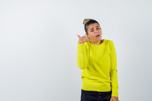 Mujer joven en suéter amarillo y pantalón negro que muestra el gesto de llamarme y parece feliz