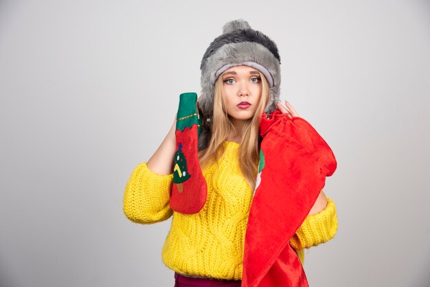 Mujer joven en suéter amarillo y sombrero sosteniendo una bolsa de Navidad roja.