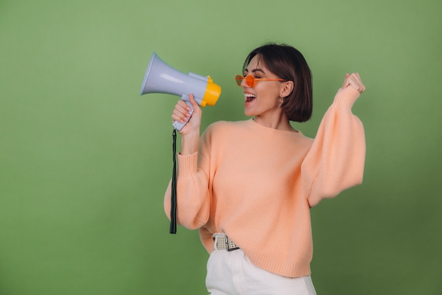 Foto gratuita mujer joven en suéter casual de anteojos de melocotón y naranja aislado en la pared verde oliva feliz gritando en el espacio de copia de megáfono