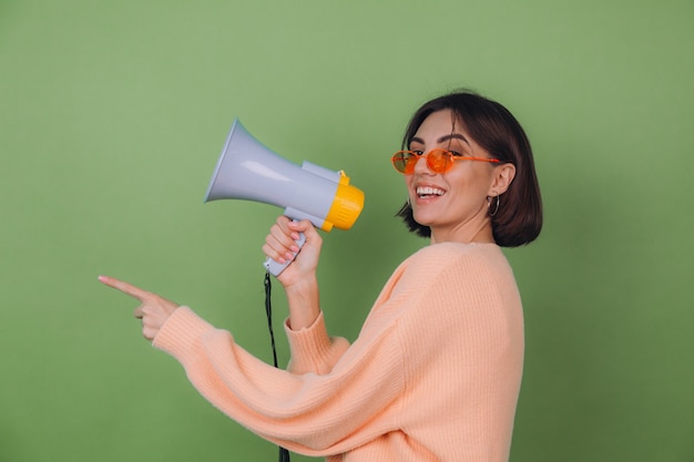 Foto gratuita mujer joven en suéter casual de anteojos naranja y melocotón aislado en la pared verde oliva feliz gritando en megáfono y apunta a la izquierda con el espacio de la copia del dedo índice