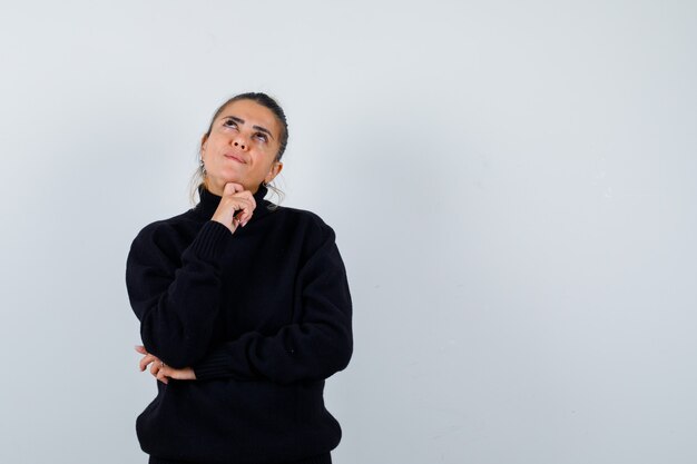 Mujer joven en suéter de cuello alto negro de pie en pose de pensamiento y mirando pensativo, vista frontal.