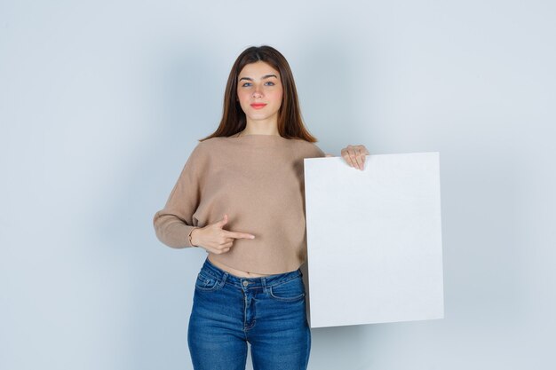 Mujer joven en suéter, jeans apuntando hacia un lado, manteniendo el tablero de papel y mirando confiado, vista frontal.