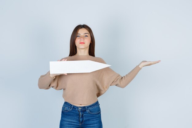 Mujer joven en suéter, jeans extendiendo el brazo, sosteniendo un cartel de papel y mirando complacido, vista frontal.