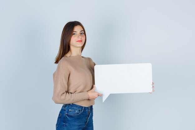 Mujer joven en suéter, jeans mostrando bocadillo de diálogo de papel, de pie de lado y mirando confiado.