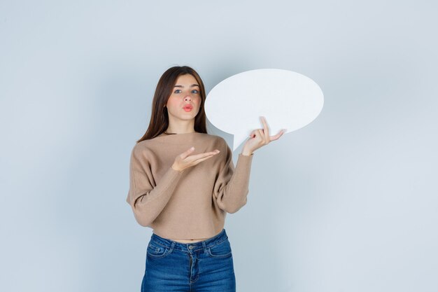 Mujer joven en suéter, jeans mostrando bocadillo de papel y mirando pensativo, vista frontal.