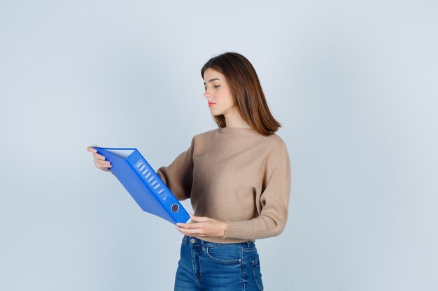 Mujer joven en suéter, jeans sosteniendo una carpeta, de pie hacia los lados y mirando pensativo.