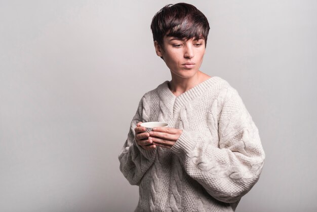 Mujer joven en el suéter de lana que sostiene la taza de café contra fondo gris