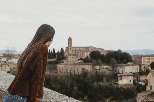 Mujer joven en un suéter marrón de pie sobre un puente y disfrutando de una hermosa vista del paisaje urbano