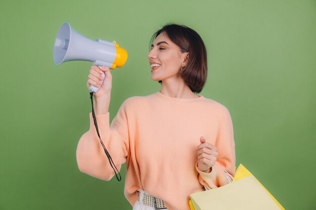 Mujer joven en suéter de melocotón casual aislado en la pared de color verde oliva grita en megáfono sosteniendo bolsas de compras, anuncia promoción de venta de descuentos