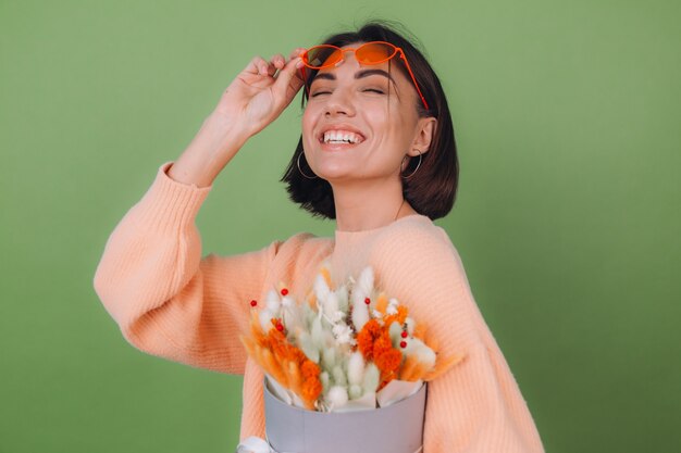 Mujer joven en suéter de melocotón casual aislado en la pared de olivo verde sostenga la composición de la caja de flor blanca naranja de flores de algodón, gypsophila, trigo y lagurus para un regalo feliz sorprendido