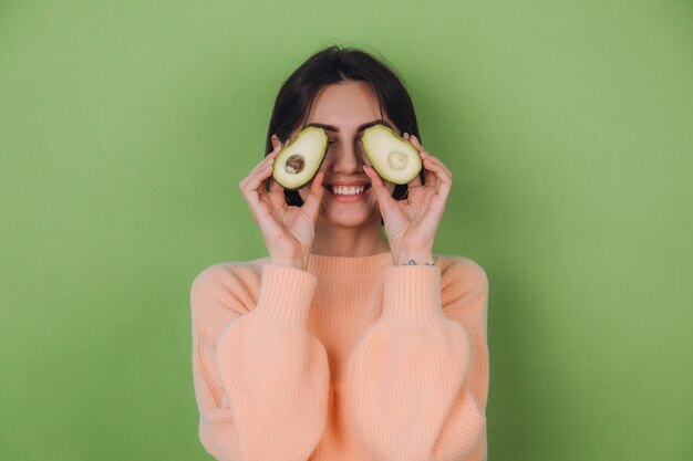 Mujer joven en suéter de melocotón casual aislado en la pared verde oliva con aguacate, concepto de salud y cuidado de la piel, espacio de copia