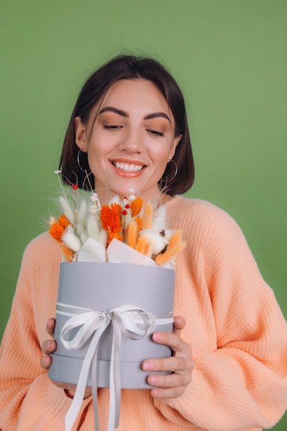 Mujer joven en suéter de melocotón casual aislado en la pared verde oliva sostenga la composición de la caja de flor blanca naranja de flores de algodón, trigo de gypsophila y lagurus para un regalo feliz sorprendido