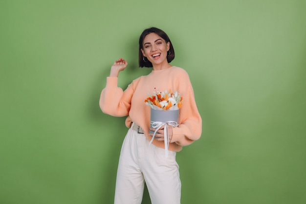 Mujer joven en suéter de melocotón casual aislado en la pared verde oliva sostenga la composición de la caja de flor blanca naranja de flores de algodón, trigo de gypsophila y lagurus para un regalo feliz sorprendido