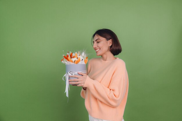 Mujer joven en suéter de melocotón casual aislado en la pared verde oliva sostenga la composición de la caja de flor blanca naranja de flores de algodón, trigo de gypsophila y lagurus para un regalo feliz sorprendido