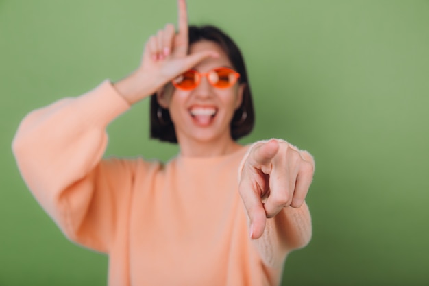 Mujer joven en suéter de melocotón casual y gafas naranjas aisladas en la pared de olivo verde apuntando a ti día de tonto de burla mostrando símbolo de cuerno copia espacio