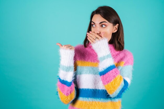 Mujer joven en un suéter multicolor brillante en azul sorprendido cubre su boca con sus palmas