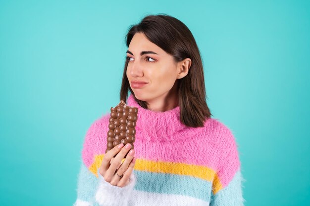 Mujer joven en un suéter multicolor brillante sobre azul sufre, llora, sostiene una barra de chocolate con leche