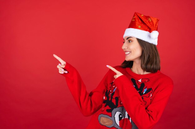 Mujer joven en suéter de navidad y gorro de Papá Noel sonriendo con entusiasmo, apuntando con su dedo hacia la izquierda a un espacio vacío