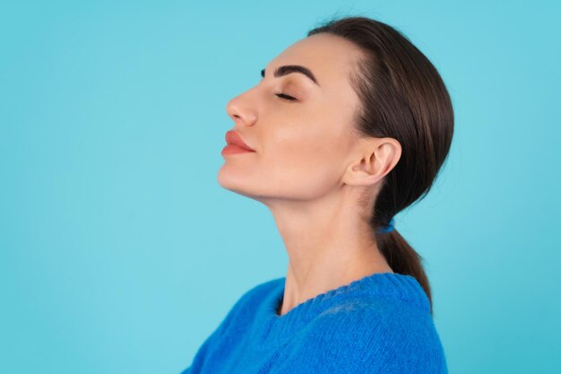 Mujer joven con un suéter de punto azul y maquillaje natural durante el día sobre un fondo turquesa, labios regordetes con lápiz labial mate desnudo