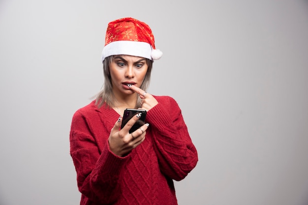 Mujer joven en suéter rojo mirando celular sobre fondo gris.