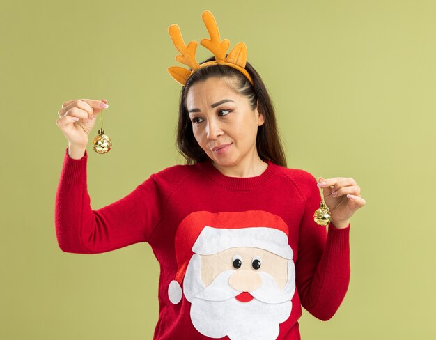 Mujer joven en suéter rojo de Navidad con borde divertido con cuernos de ciervo sosteniendo bolas de Navidad mirando bola con sonrisa en la cara tratando de hacer una elección de pie sobre la pared verde