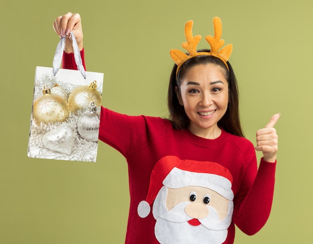 Mujer joven en suéter rojo de Navidad con borde divertido con cuernos de ciervo sosteniendo una bolsa de papel con regalo de Navidad con una sonrisa en la cara mostrando los pulgares para arriba de pie sobre la pared verde