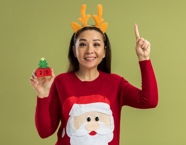 Mujer joven en suéter rojo de Navidad con borde divertido con cuernos de ciervo sosteniendo cubos de juguete con fecha veinticinco mirando a la cámara feliz y alegre mostrando el dedo índice de pie sobre fondo verde