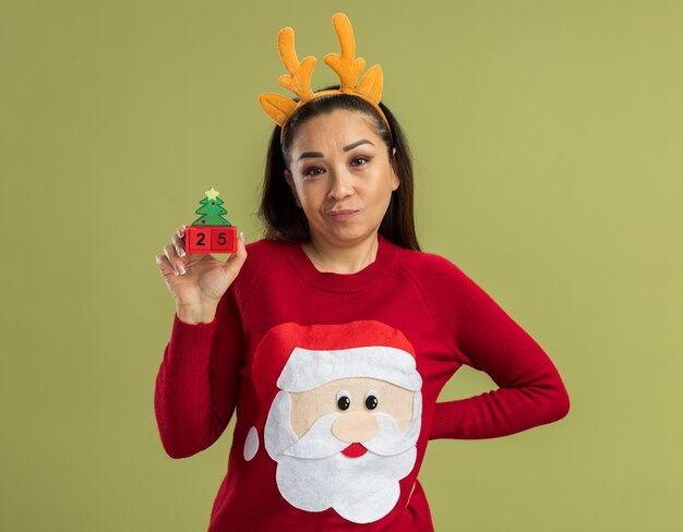 Mujer joven en suéter rojo de Navidad con borde divertido con cuernos de venado mostrando cubos de juguete con fecha veinticinco con sonrisa escéptica de pie sobre la pared verde