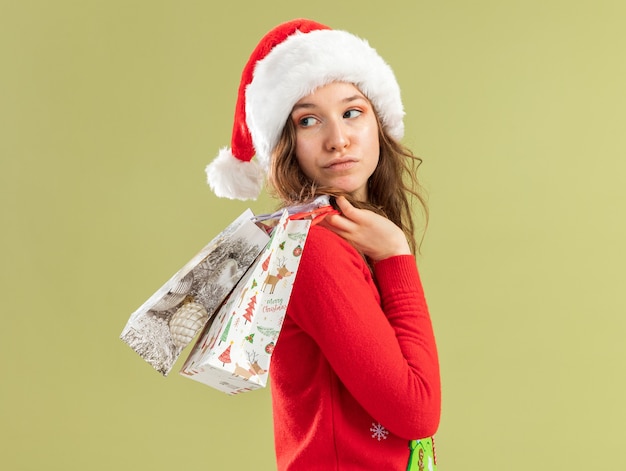 Mujer joven en suéter rojo de Navidad y gorro de Papá Noel sosteniendo bolsas de papel con regalos de Navidad mirando hacia atrás con expresión de confianza