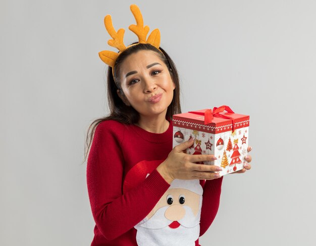 Mujer joven en suéter rojo de Navidad vistiendo borde divertido con cuernos de ciervo sosteniendo regalo de Navidad mirando con sonrisa escéptica