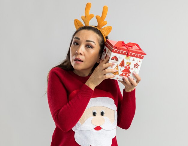 Mujer joven en suéter rojo de Navidad vistiendo un borde divertido con cuernos de ciervo sosteniendo un regalo de Navidad sobre su oreja tratando de escuchar algo