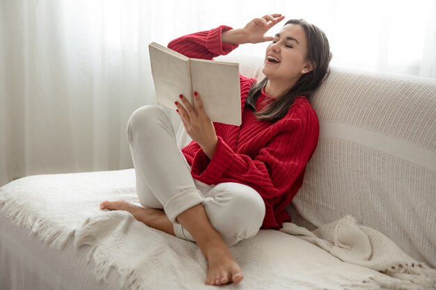 Mujer joven con un suéter rojo en el sofá en casa con un libro en sus manos.