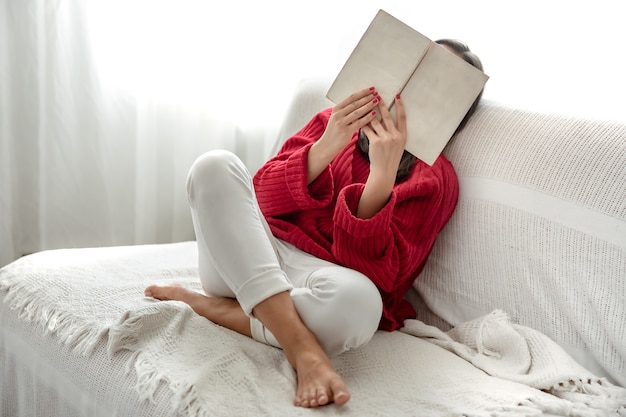 Mujer joven con un suéter rojo en el sofá en casa con un libro en sus manos.