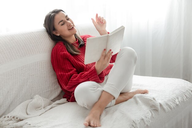 Mujer joven con un suéter rojo en el sofá en casa con un libro en sus manos.