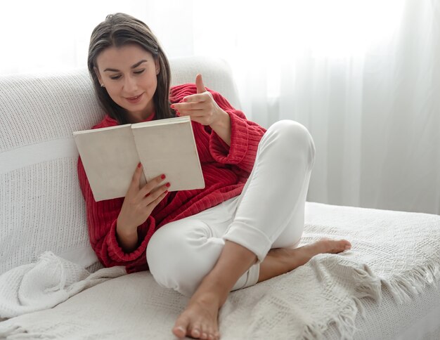 Mujer joven con un suéter rojo en el sofá en casa con un libro en sus manos.