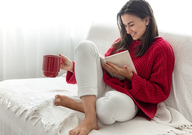Mujer joven con un suéter rojo con una taza roja lee un libro.
