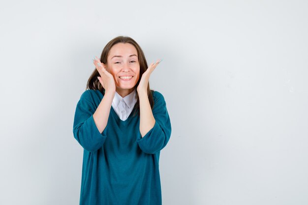 Mujer joven en suéter sobre camisa blanca manteniendo las manos cerca de la cara y mirando alegre, vista frontal.